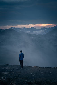 Rear view of man standing on mountain against cloudy sky during sunset