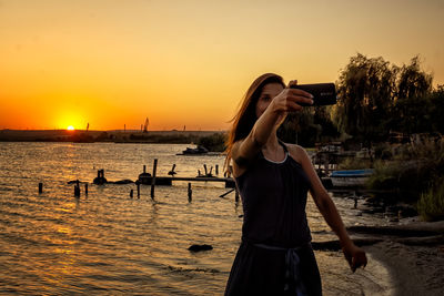 Woman taking selfie on mobile phone while standing by sea against sky during sunset