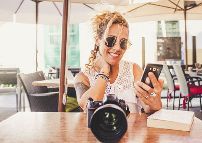 Smiling woman holding smart phone while sitting with camera at table