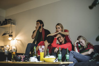 Disappointed fans watching soccer match while sitting on sofa at home