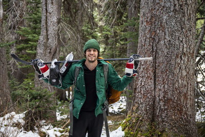 Cheerful young man with hockey stick and ice skates on shoulders smiling and looking at camera while standing near trees in snowy forest
