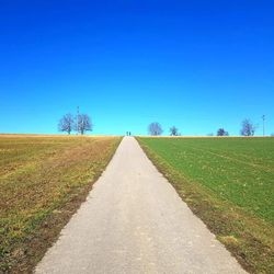 Empty road amidst field against clear blue sky