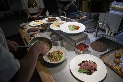 High angle of crop anonymous chef adding fried chicken fillets into plate with pasta while preparing food with colleagues in modern restaurant kitchen