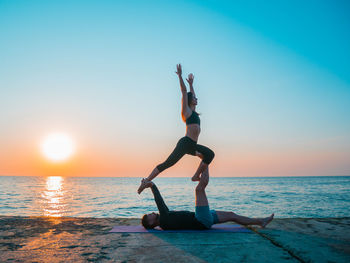 Side view of couple practicing yoga on pier by sea against sky during sunset