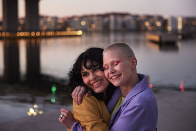 Happy young female friends or gay couple hugging, river in background