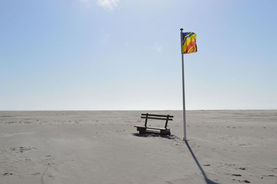Flag by empty bench on sandy field against clear sky