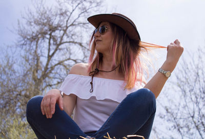 Woman in sunglasses and hat sitting against bare tree