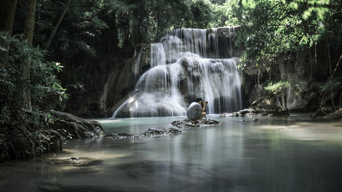 Scenic view of waterfall with man crouching on rock in forest