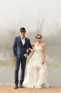Portrait of smiling bride and groom wearing sunglasses while standing against lake during foggy weather
