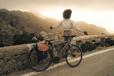 Woman with arms outstretched looking at mountains while standing roadside with cycle on vacation