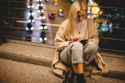 Young woman using smart phone while sitting on bridge in city at night