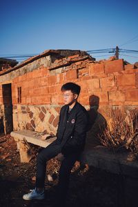 Young man sitting on brick wall against sky