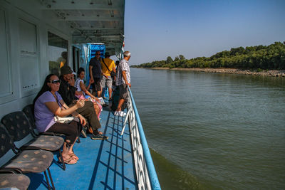 People sitting on riverbank against clear sky