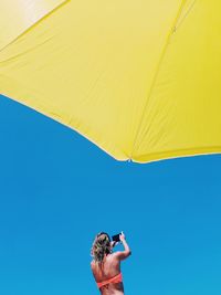 Rear view of woman photographing clear sky through mobile phone at beach