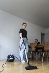 Young woman cleaning hardwood floor with vacuum cleaner near girlfriend using laptop at home