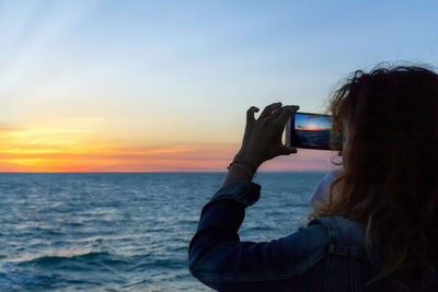 Rear view of person photographing sea against sky during sunset