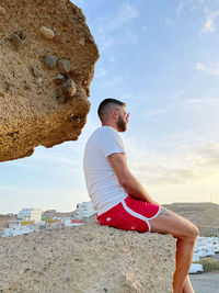 Side view of young man on rock at beach against sky