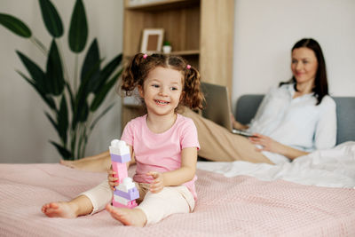 A cute girl is playing with a constructor on the bed while her mom is working on a laptop