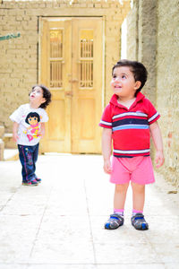 Full length of cute smiling siblings looking away while standing outside house