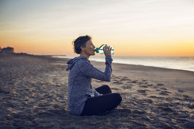 Woman drinking water while sitting at beach against sky