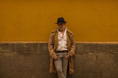 Portrait of adult man in hat and coat against yellow wall on street