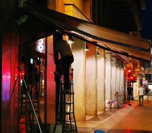 Rear view of man standing on illuminated street against buildings at night