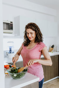 Female in casual clothes standing at kitchen table and mixing fresh vegetables in salad in glass bowl in daytime