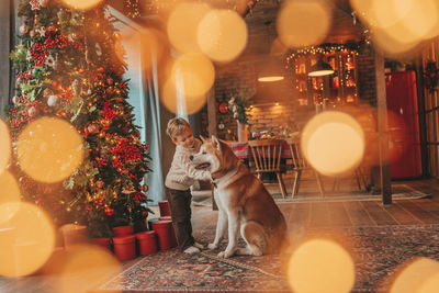 Candid authentic happy little boy in knitted beige sweater hugs dog with bow tie at home on xmas