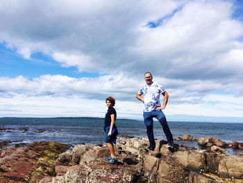 Father and son standing on rock at beach against sky