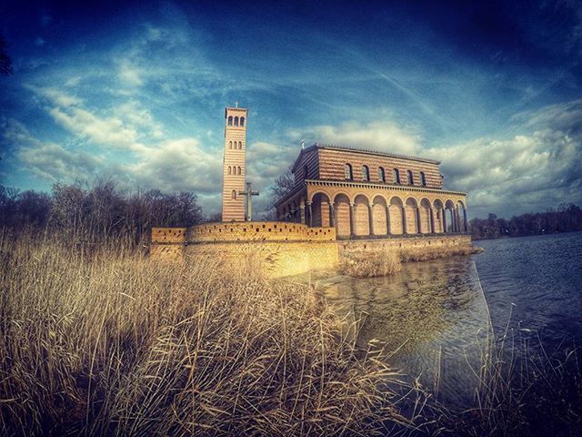 architecture, built structure, water, sky, building exterior, cloud - sky, cloud, blue, grass, river, nature, tranquility, waterfront, day, tranquil scene, outdoors, lake, travel destinations, reflection, no people