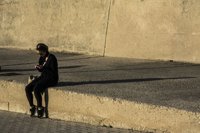 Full length of woman using mobile phone while sitting against wall
