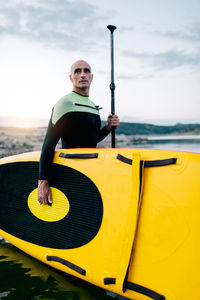 Side view of male surfer in wetsuit standing with yellow sup board and paddle in sea water