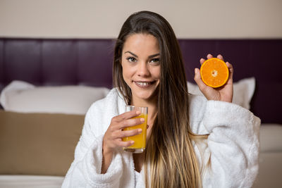 Portrait of smiling beautiful woman holding drink and fruit while sitting on bed at hotel