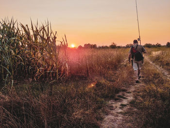 Rear view of man carrying fishing rod on field against sky during sunset