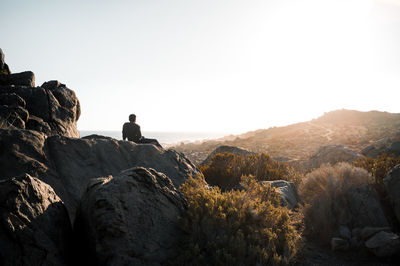 Rear view of man sitting on rock against clear sky at sunset