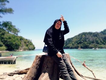 Portrait of woman sitting on tree stump by sea against sky