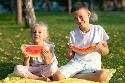 Brother and sister holding watermelon while sitting in park