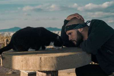 Side view of mid adult man looking at cat on table against sky