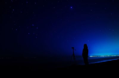 Silhouette man standing with tripod on shore at beach against blue sky