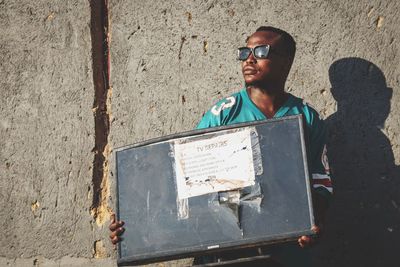 Low angle view of man standing against wall holding a broken television. 