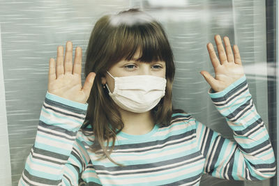 A sad dark-haired teenager girl with a protective mask stands with her hands pressed to the glass 