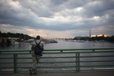 Rear view of man standing on railing by river against sky