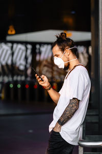 Side view of man wearing mask using mobile phone while standing outdoors