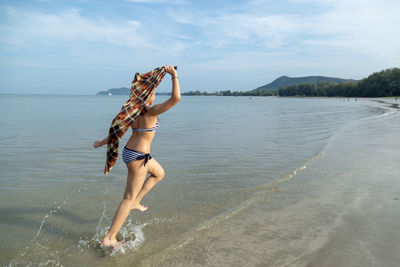 Teenage girl wearing bikini running at shore against sky