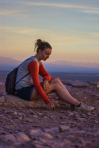 Side view of woman sitting rock against sky during sunset