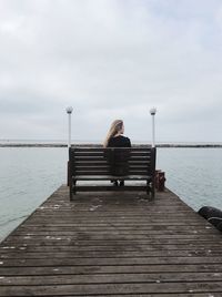 Rear view of man sitting on pier over sea against cloudy sky