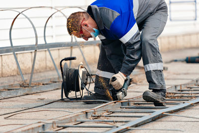 Builder in overalls leans over and cuts off metal sheet with angle grinder and sparks fly. working