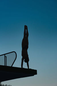 Low angle view of silhouette mid adult woman doing handstand on diving platform against clear sky during sunset