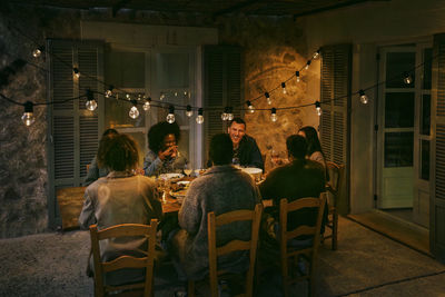 Happy multiracial female and male friends sitting at dining table in patio during dinner party at night