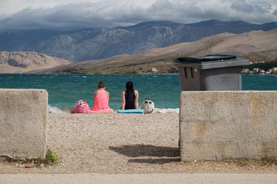 Rear view of women sitting at beach against cloudy sky
